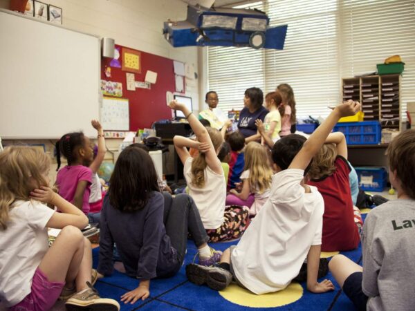 Young students sitting on a classroom floor raising their hands