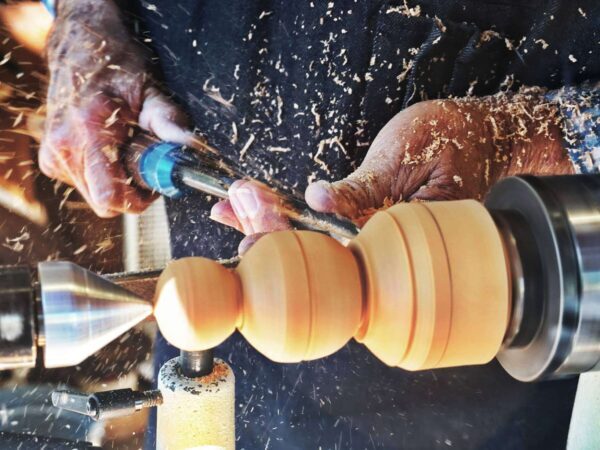 Hands shaping a piece of wood on a lathe, with shavings flying