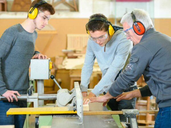 Instructor and two students in ear defenders and safety glasses working at a woodworking machine