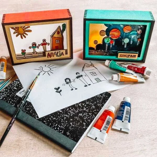 Laser-cut wooden picture frames holding drawings, beside paints and a notebook