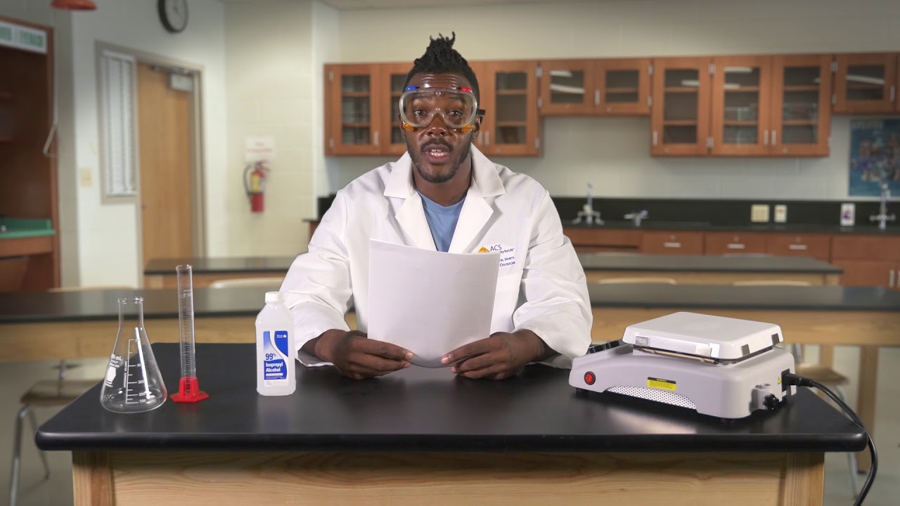 Person in a white lab coat and goggles sits at a lab bench holding a paper, with lab equipment (beaker, graduated cylinder, hand sanitizer) and a hot plate on the counter behind him.